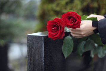 Woman with red roses near black granite tombstone outdoors, closeup. Funeral ceremony