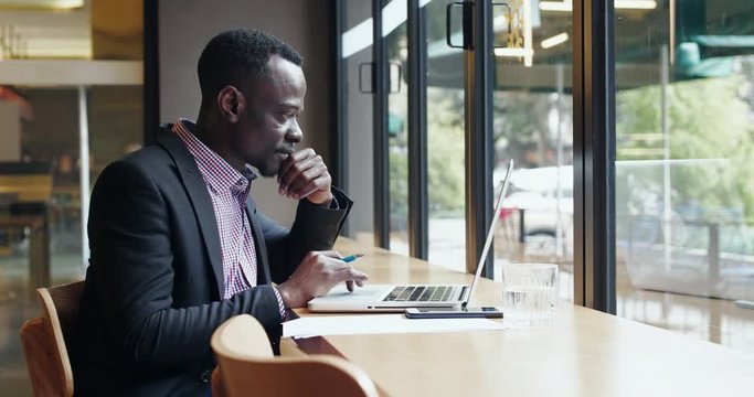 Side View Of Young African Businessman Working In The Office Sitting By The Window Using Laptop Black Freelancer Hand On Chine Thinking Busy Focus Looking At Computer Screen 