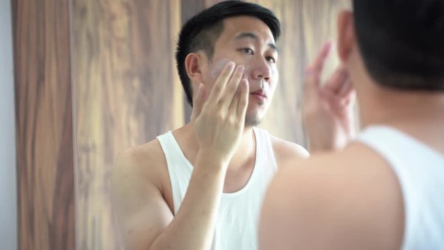 Back view of Asian male in white shirt using a skincare cream for face in front of mirror in bathroom