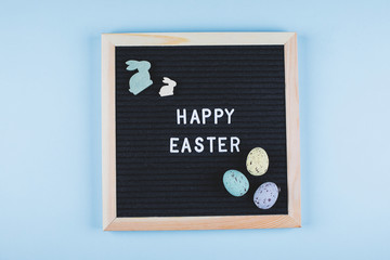 Easter Card Flat Lay Concept. Greeting board with text Happy Easter and pastel colorful eggs, wooden bunny on blue background. Top view.