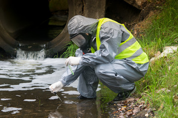 Fototapeta na wymiar a scientist, in a protective suit, mask and gloves, takes a liquid from the river in test tubes, for a sample, against the background of a drainpipe