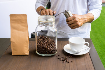Person grinding coffee with manual coffee grinder beside blur of Blank brown paper bag foreground - obrazy, fototapety, plakaty
