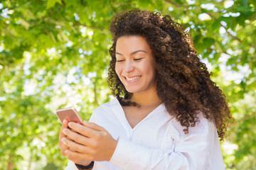 Smiling curly lady looking at smartphone in spring park. Low angle shot of cheerful brunette woman using modern phone. Technology concept