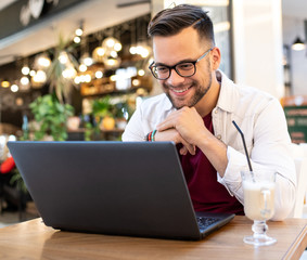 A young man in a cafe drinking a beverage and using his laptop.