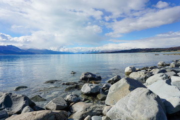 The sandy beach by the lake, behind the mountains, there are rocks and the sky.