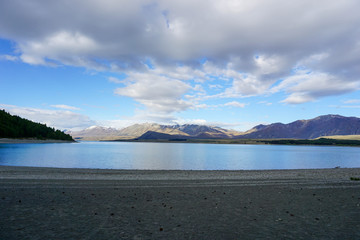 The sandy beach by the lake, behind the mountains, there are rocks and the sky.