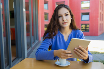 Calm young woman holding tablet and looking at camera. Front view of tranquil lady sitting at table at outdoor cafe. Technology concept