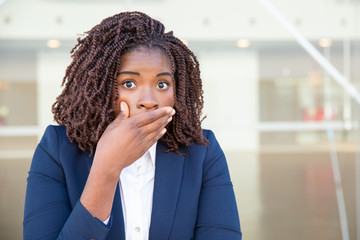 Fototapeta na wymiar Shocked young businesswoman closing mouth. Scared young African American businesswoman holding hand on face and looking at camera. Shock concept