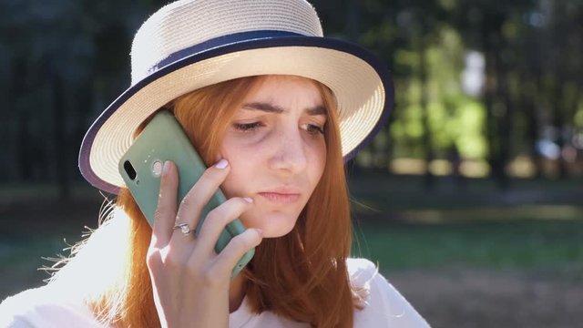 Portrait of angry pissed off teenage girl with red hair wearing straw hat and pink earphones talking fiercely on mobile phone.