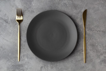 Empty gray plate with a gold knife and fork on a gray concrete background