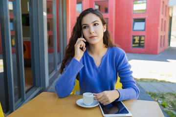 Calm young woman sitting at outdoor cafe and talking on phone. Front view of tranquil lady sitting at table with tablet and cup of coffee. Technology and communication concept