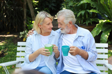 caucasian elderly couple with blue shirt and blue jean sitting and drinking coffee in garden during summer time on wedding anniversary day