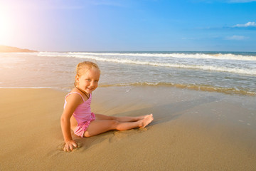 Little pretty Girl in pink swimsuit sitting on the beach in water waves and looking at the morning sea and sky