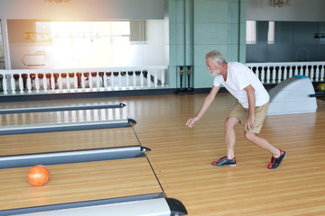 Indoor shot attractive bearded senior caucasian man in white shirt playing bowling in sport club in hotel with happy face. Senior people life style concept