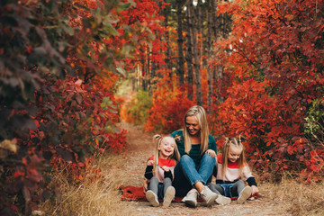 Blonde mother and her twin daughters are walking in the autumn forest among red trees.