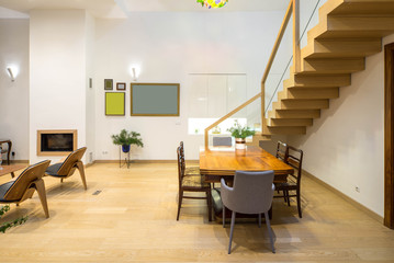 Light interior of room in private house. Wooden staircase, table and chairs. Chandelier. Black fireplace.