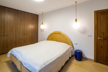 Light interior of bedroom. Wooden furniture. Closed door. Round lamp. Made-up bed with white cover.