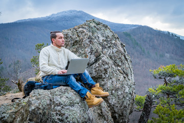 Hipster man working with notebook sitting on the rocky mountain on beautiful scenic clif background