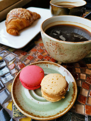 Two ceramic cups of black coffee with croissants and macaroni cakes on a vintage table