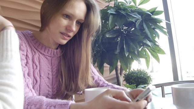 Two pretty women friends are looking pictures on social media in smartphone and conversating it on coffee break in cafe interior with decorative tree on background. Closeup portrait view shot 4K video