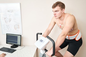 Fototapeta na wymiar Man patient, pedaling on a bicycle ergometer stress test system for the function of heart checked. Athlete does a cardiac stress test in a medical study, monitored by the doctor.