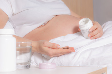 Obraz na płótnie Canvas Unrecognizable Pregnant Woman in bed holding vitamins, pills in hand before intake. Glass of water is on bedside table. Pregnancy healthy lifestyle. Snow-white bedding.
