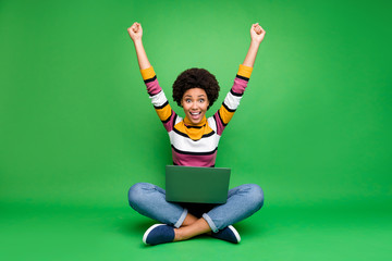 Full length photo of crazy funky afro american girl work laptop sit legs crossed win coworking deal scream wow omg raise fists wear denim jeans shine outfit isolated green color background - obrazy, fototapety, plakaty