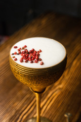 Cocktail with white foam decorated with red dried peppers served in the metal gold glass on the wooden bar table
