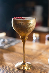 Cocktail with white foam decorated with red dried peppers served in the metal gold glass on the wooden bar table