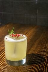Green lime cocktail with white foam served in rocks glass on the wooden bar table decorated with red dried pepper and rosemary