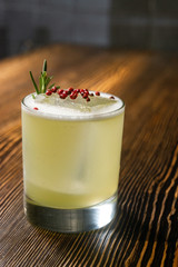 Green lime cocktail with white foam served in rocks glass on the wooden bar table decorated with red dried pepper and rosemary