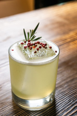 Green lime cocktail with white foam served in rocks glass on the wooden bar table decorated with red dried pepper and rosemary
