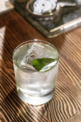 Transparent cocktail in rocks glass decorated with one big ice cube and green leaf served on the wooden bar desk.