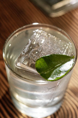 Transparent cocktail in rocks glass decorated with one big ice cube and green leaf served on the wooden bar desk.