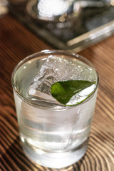 Transparent cocktail in rocks glass decorated with one big ice cube and green leaf served on the wooden bar desk.