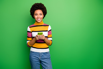 Photo of cheerful blogger dark skin wavy lady holding telephone hands modern technology addicted user person wear casual striped jumper jeans isolated green color background