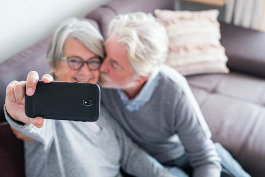 cute couple of two seniors married taking a selfie together sitting on the sofa at home - mature man kissing his wife while she's taking a photo