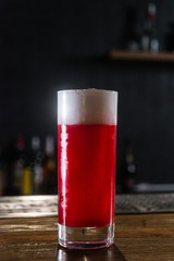 A single-colored red transparent cocktail, refreshing in a tall glass with white foam and violet flower on it served in tall glass on the wooden bar desk