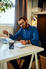 Fototapeta premium Young creative caucasian bearded environmentalist with curly hair sitting in his office, touching windmill model and drawing new better model in notebook.