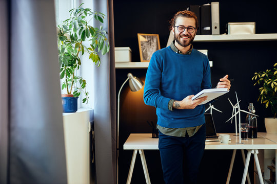 Young smiling caucasian innovative creative graphic designer standing in his office and writing ideas in notebook.