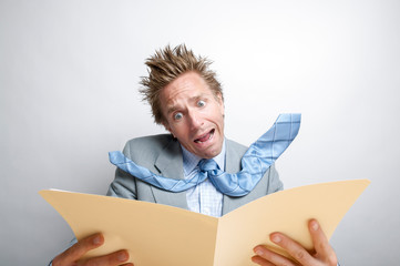 Shocked businessman with tie flying opening a file folder full of bad news at his desk