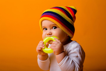 baby boy in a white bodysuit and multicolored hat holding a toy, sitting on a yellow background,...