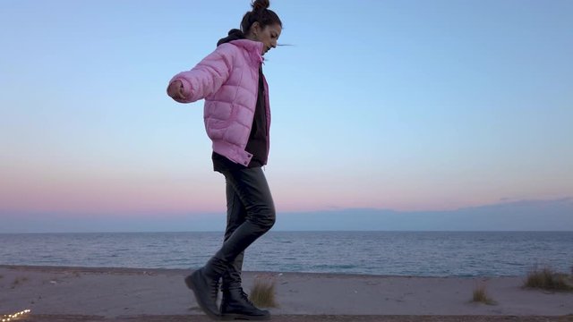 Giovane ragazza con giubbotto rosa sta facendo l'equilibrista su un tronco nella spiaggia vicino al mare al tramonto.