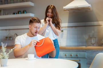 young beautiful woman greeting her surprised husband with present in red bag on saint valentine's day during breakfast in the kitchen, happy romantic unniversary