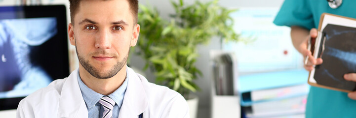 Fototapeta premium Portrait of handsome doctor working in modern clinic with colleagues. Young physician in white lab coat and blue shirt looking in camera with joy. Medical treatment concept