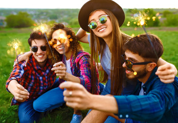 Group of friends having fun with sparklers sitting on grass in summer park. Teenagers having fun...