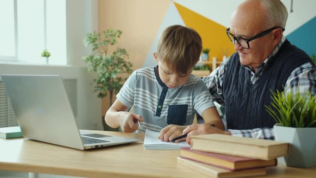 Old man and child are using laptop computer doing homework then doing high-five smiling sitting at desk together. Contemporary lifestyle and technology concept.