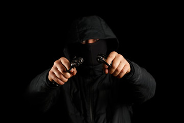 A man with two pistols on a black background. The gun is in focus, the masked man is blurred.