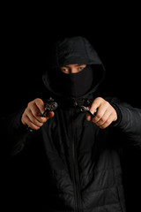 A man with two pistols on a black background. The gun is in focus, the masked man is blurred.