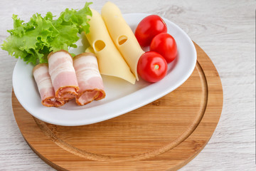 On a white dish are fresh  and tasty products for a quick snack-cheese,bacon,tomatoes and greens